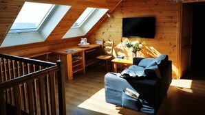 A cozy attic room with wooden walls, a TV mounted on the wall, a table with a vase of flowers, and a sofa with a blanket.