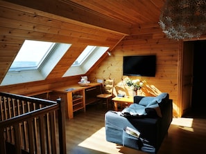 Um quarto de sótão aconchegante com paredes de madeira, uma televisão fixada na parede, uma mesa com um vaso de flores e um sofá com um cobertor.
