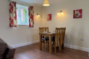 A dining area with a wooden table and chairs, a window with floral curtains, wall-mounted lights, and a painting.