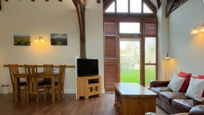A living room with a wooden dining table, chairs, a television on a wooden stand, a leather sofa with cushions, and a large window with wooden shutters. - Within the South Downs National Park, 7 miles north of Brighton. (Hassocks)