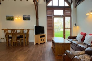 Una sala de estar con una mesa de comedor de madera, sillas, un televisor sobre un mueble de madera, un sofá de cuero con cojines y un ventanal con contraventanas de madera.
