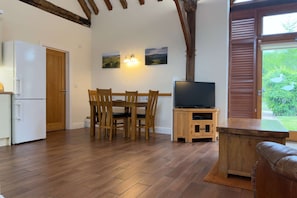 Una cocina con un refrigerador blanco, mesa de comedor de madera con sillas, un televisor sobre un mueble de madera y un ventanal con vistas a zonas verdes.