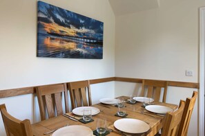 Un coin repas avec une table en bois dressée pour quatre personnes, des chaises et un tableau représentant un coucher de soleil sur un pont, accroché au mur.