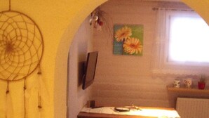 Une pièce chaleureuse avec une table en bois, un attrape-rêves accroché au mur et une fenêtre ornée d’une peinture de fleurs.
