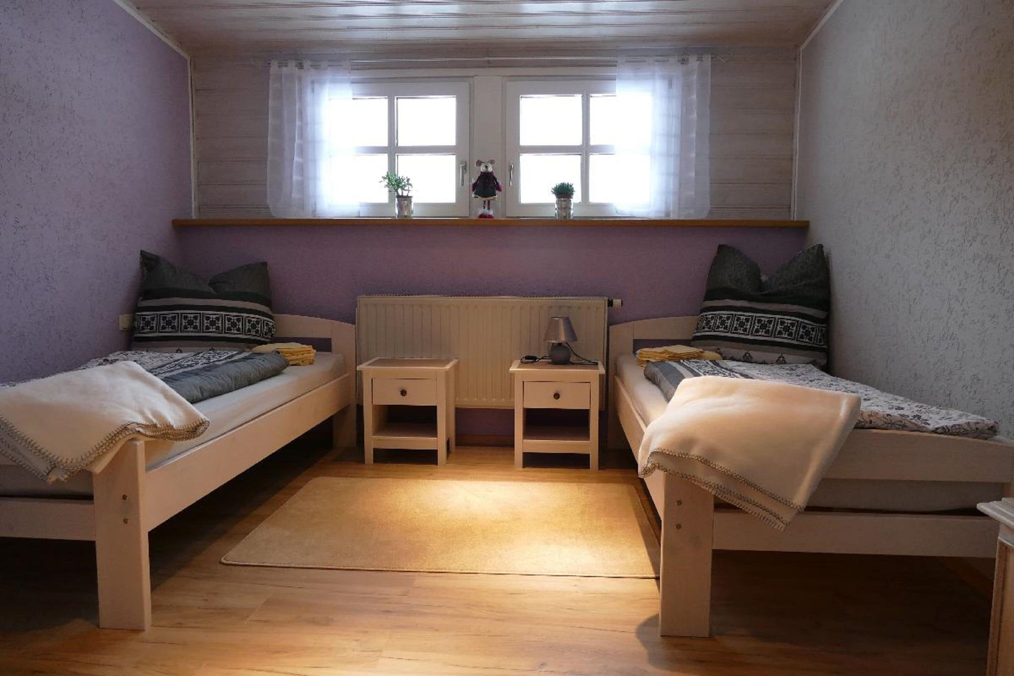 A room with two single beds, a window with curtains, a radiator, and a small table with a lamp.