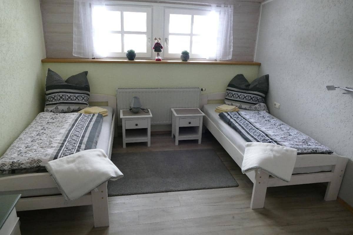 Une petite chambre propre, avec deux lits simples, une table de chevet, un radiateur et une fenêtre donnant sur un paysage agréable.