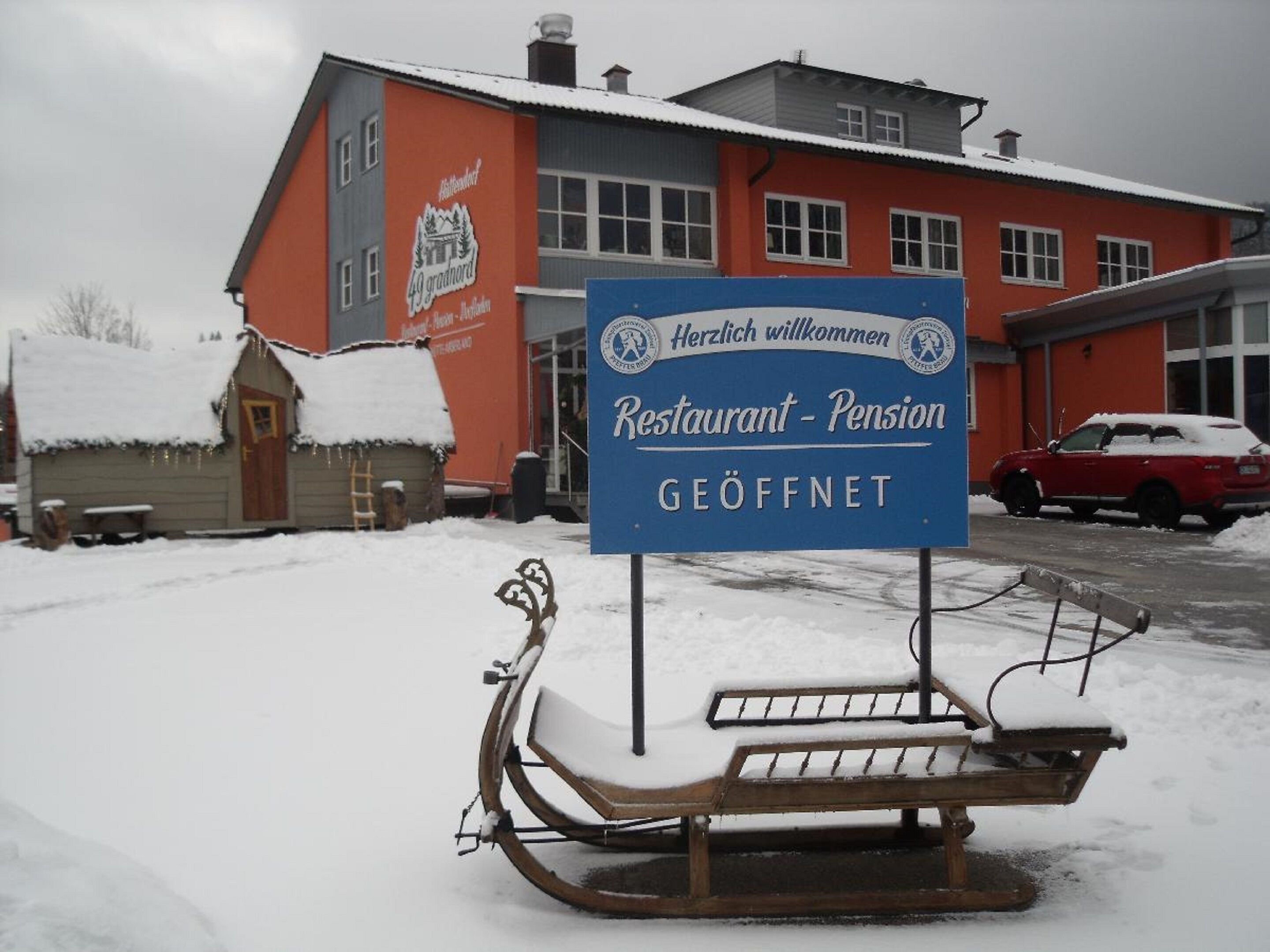 A snow-covered outdoor seating area with a bench and sled, in front of a building with a sign reading "Restaurant - Pension GEÖFFNET."