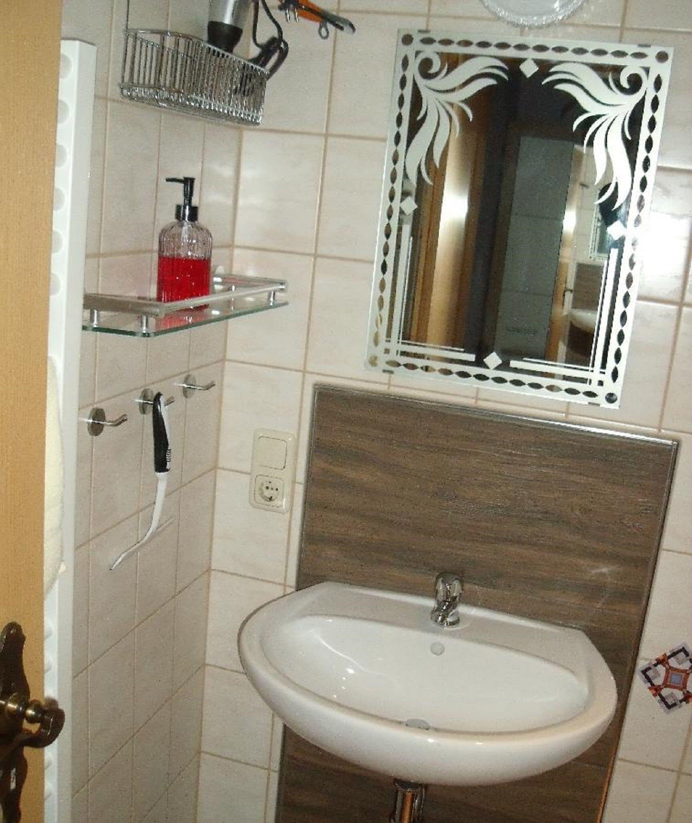 Une salle de bain avec un lavabo blanc, un miroir doté d’un cadre décoratif, un distributeur de savon posé sur une étagère en verre et un porte-serviettes.
