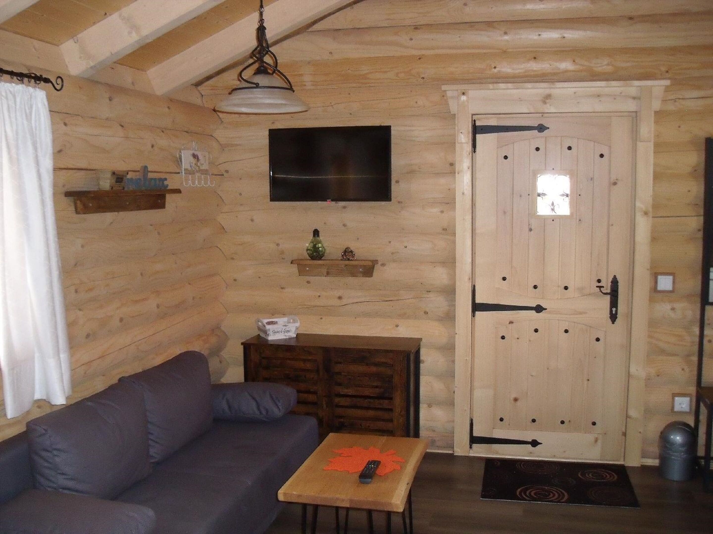 Interior acogedor de una cabaña de troncos con un sofá, una mesita, un televisor montado en la pared y una puerta de madera con un ventanal.