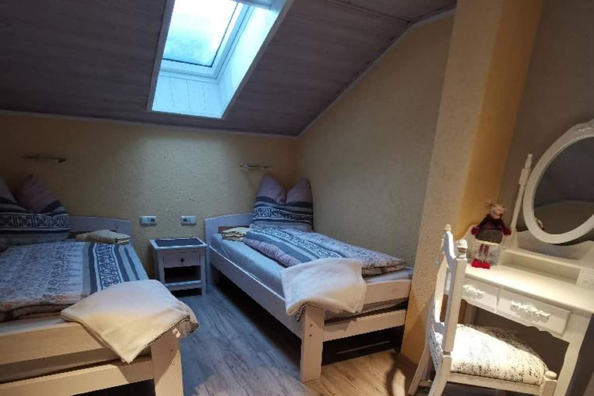 Une chambre de taille modeste, équipée de deux lits, d’une lucarne, d’une petite table et d’une coiffeuse avec un miroir.