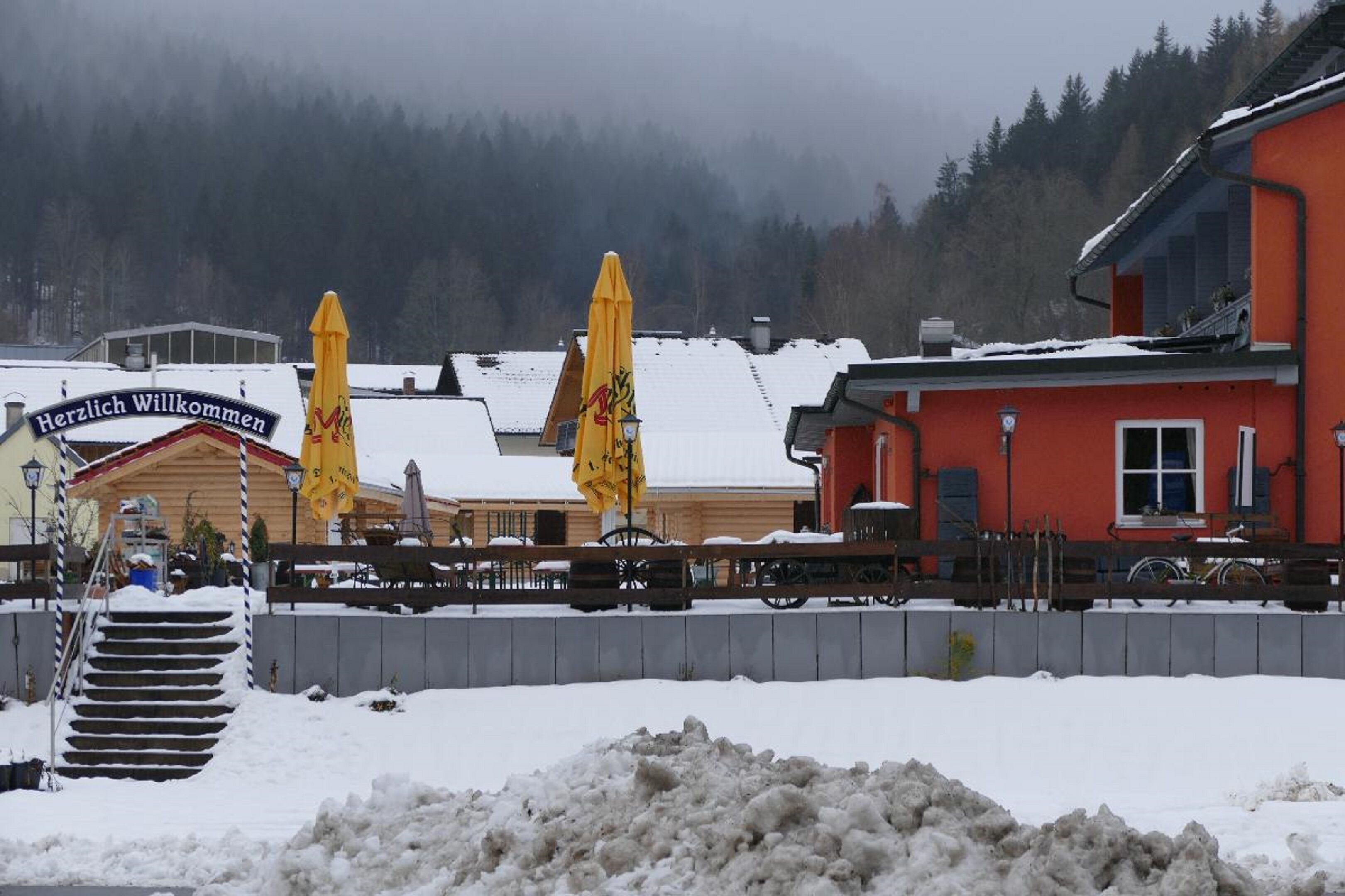 A snow-covered outdoor area with a wooden building labeled "Herzlich Willkommen" and yellow umbrellas.