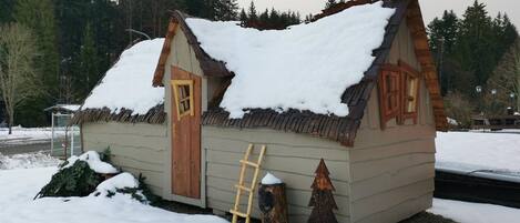 Una pequeña cabaña de madera con techo nevado y puerta, rodeada por un paisaje invernal y árboles.