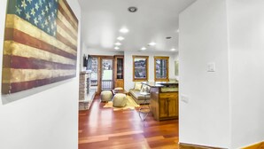 Uma sala de estar ampla com piso de madeira, uma grande pintura da bandeira americana, uma lareira de pedra e uma área de cozinha com armários de madeira e bancada.