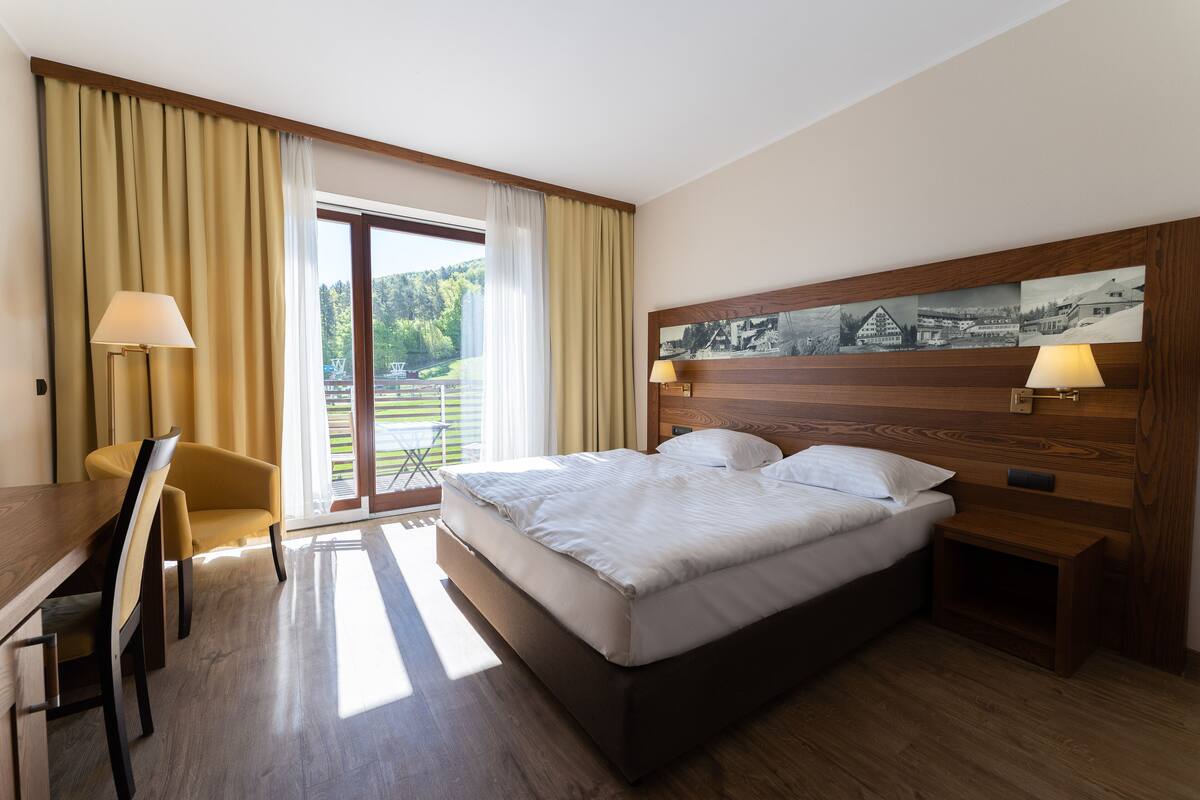 A hotel room with a bed, a desk, a chair, a nightstand, and a window with curtains.