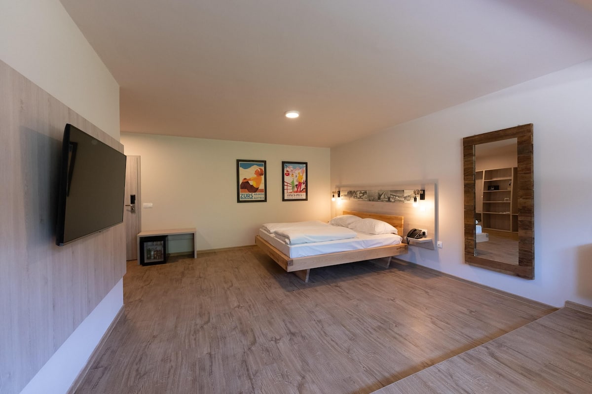A modern bedroom with a wooden floor, a bed with white linens, a mounted TV, and two framed posters on the wall.