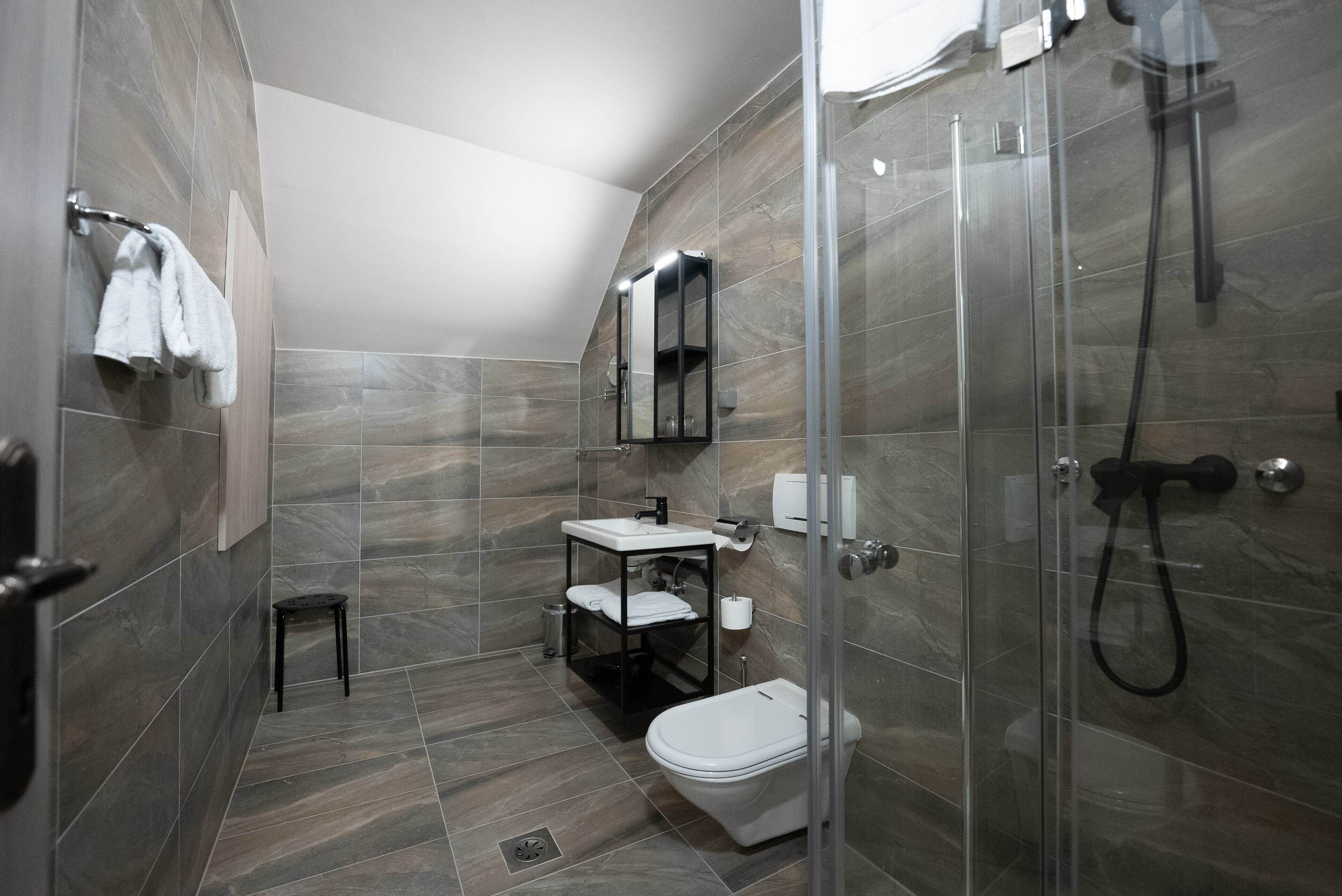 A modern bathroom with a glass-enclosed shower, a white toilet, a sink with a mirror, and a small stool.