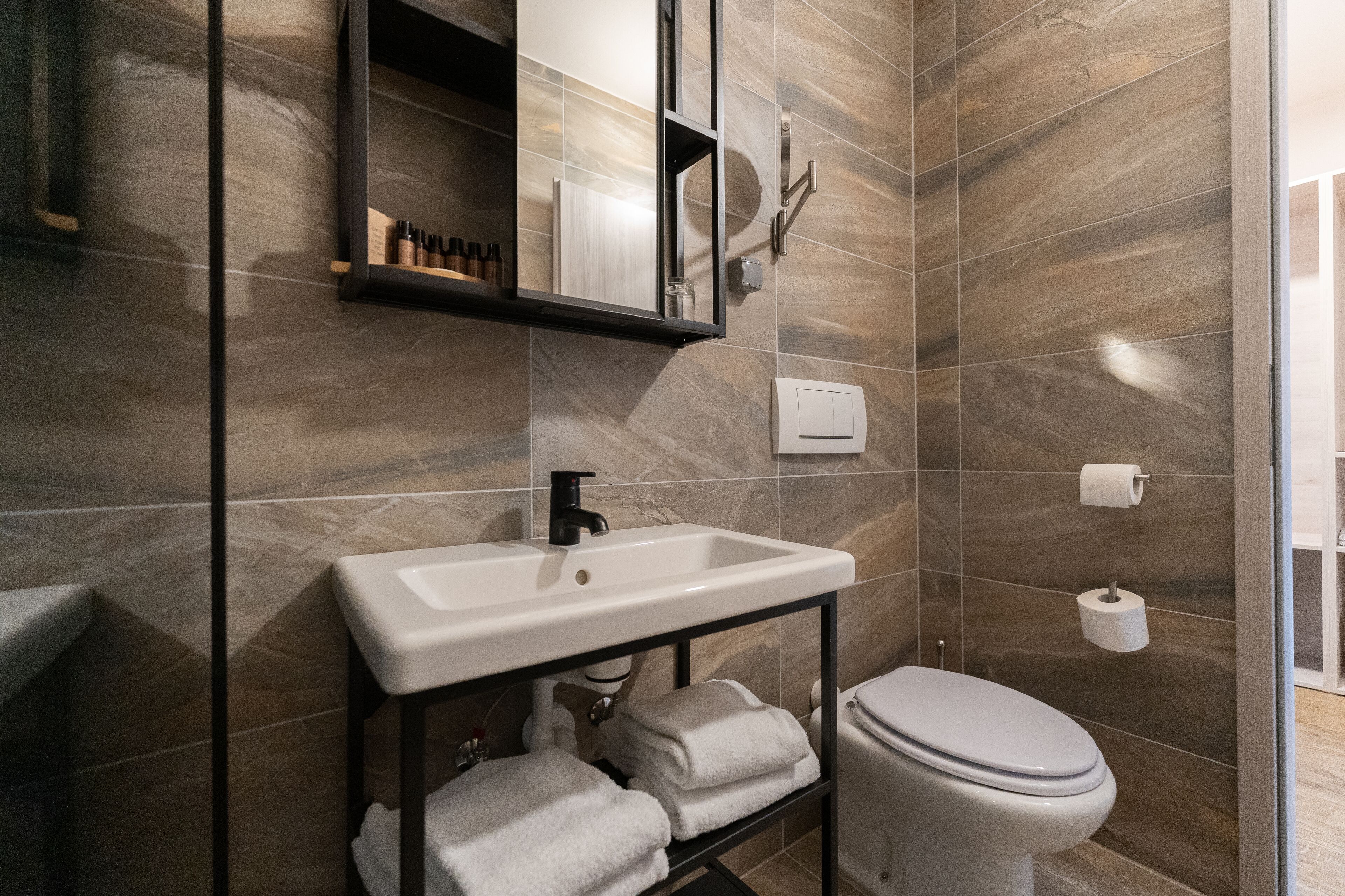 Ein modernes Badezimmer mit einem weißen Waschbecken, einem schwarzen Wasserhahn und einer Toilette. Ein Handtuchhalter mit gefalteten Handtüchern und ein Spiegel über dem Waschbecken.