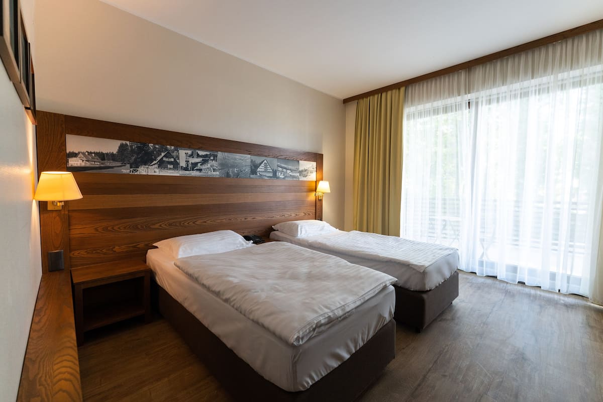 A hotel room with two beds, wooden headboard, large windows with sheer curtains, and a wall-mounted picture.
