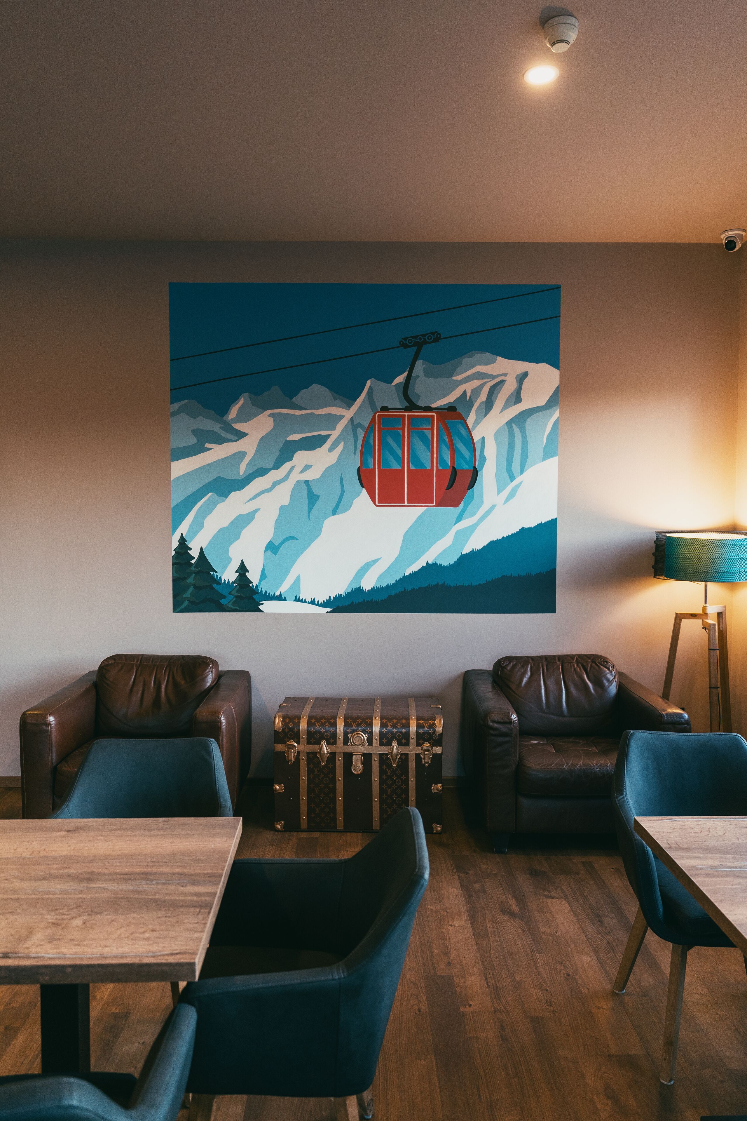 Una habitación con una mesa de madera, dos sillas azules, un sofá de cuero marrón y un cuadro colgado en la pared que muestra una escena de montaña con un teleférico rojo.