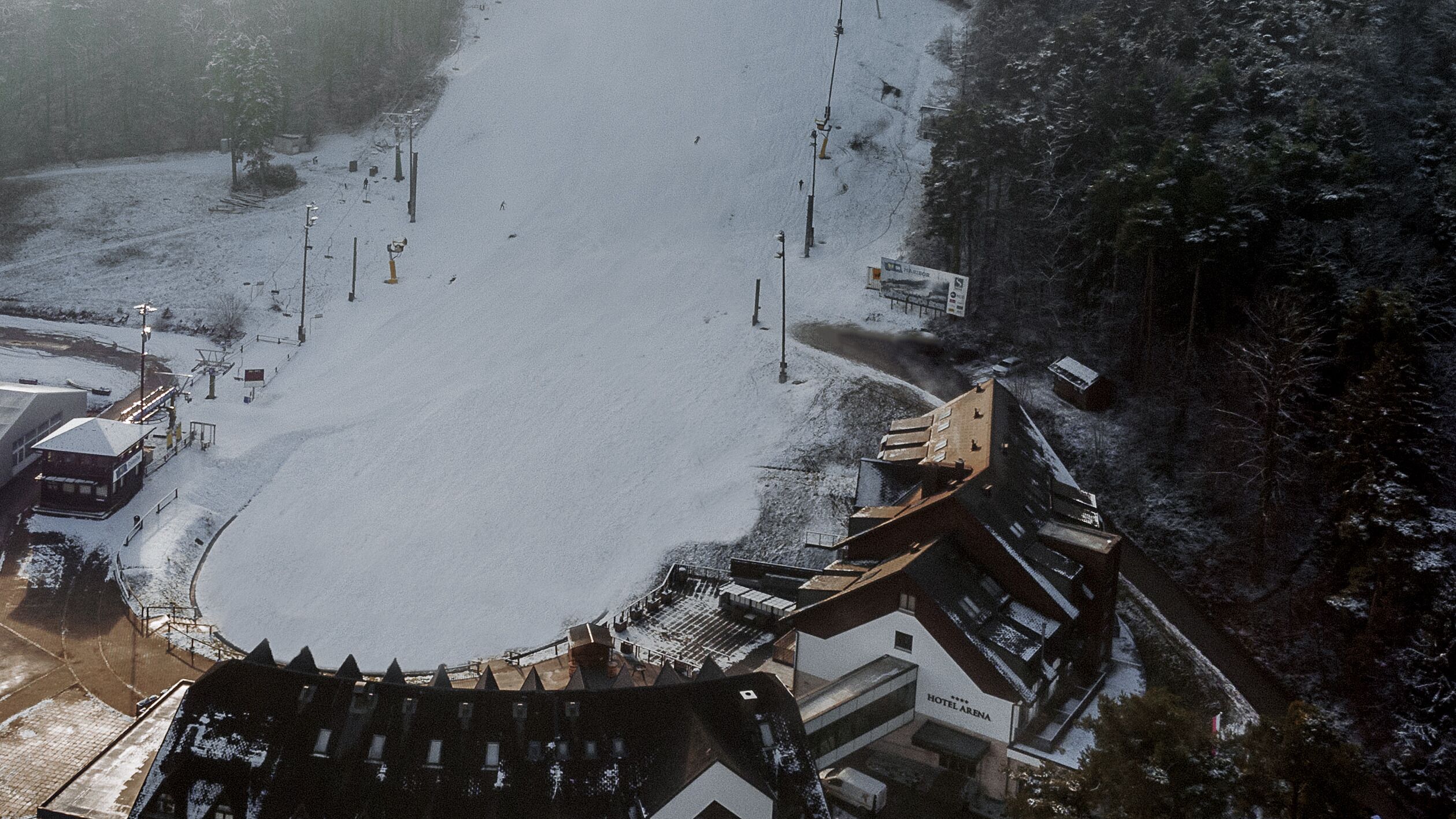 Vista aérea de um hotel com um pátio coberto de neve, um teleférico de esqui e área florestal ao redor.