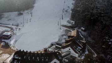 Vista aérea de um hotel com um pátio coberto de neve, um teleférico de esqui e área florestal ao redor.