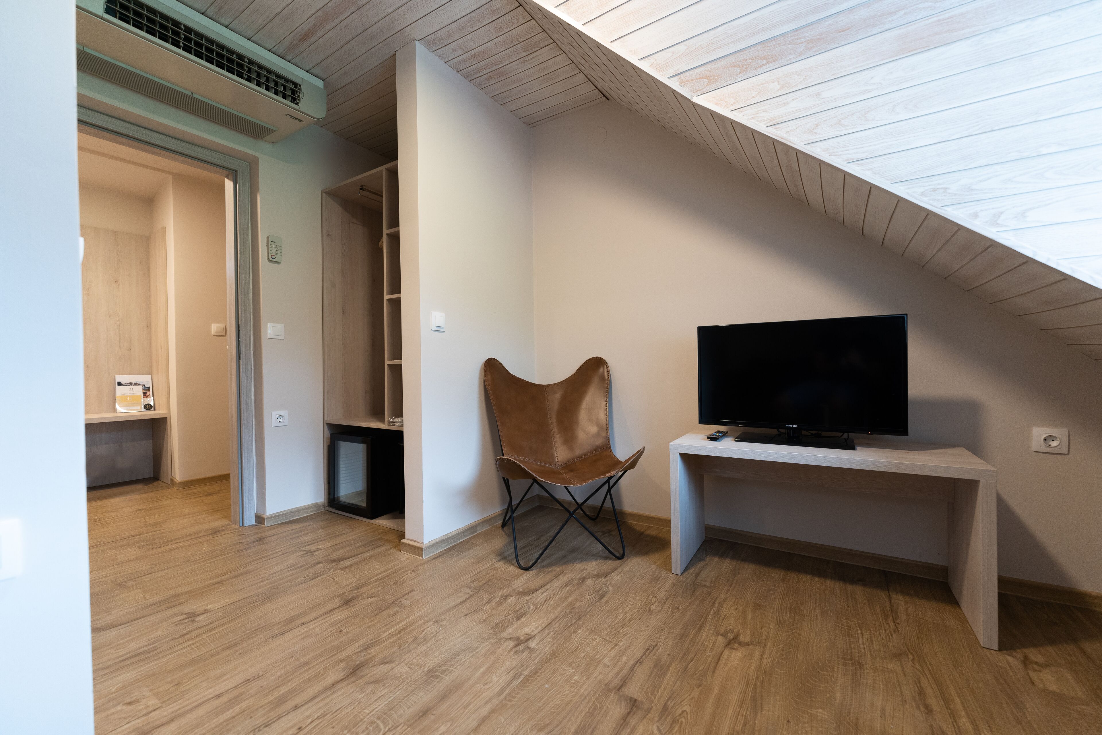 Una camera moderna con soffitto spiovente, una televisione su un supporto bianco e una poltrona in pelle marrone.