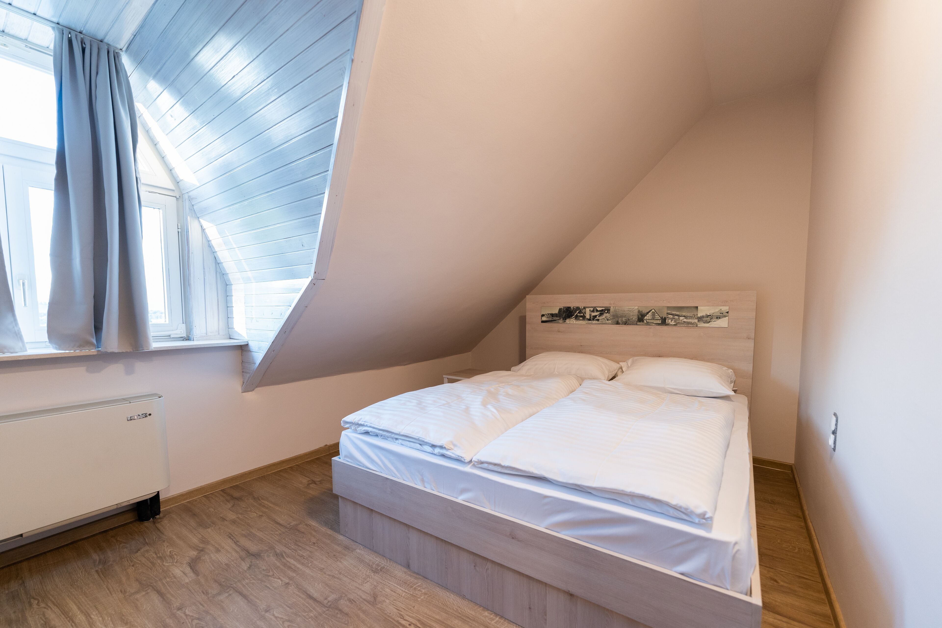 A bedroom with a sloped ceiling, a bed with white bedding, a radiator, and a window with curtains.