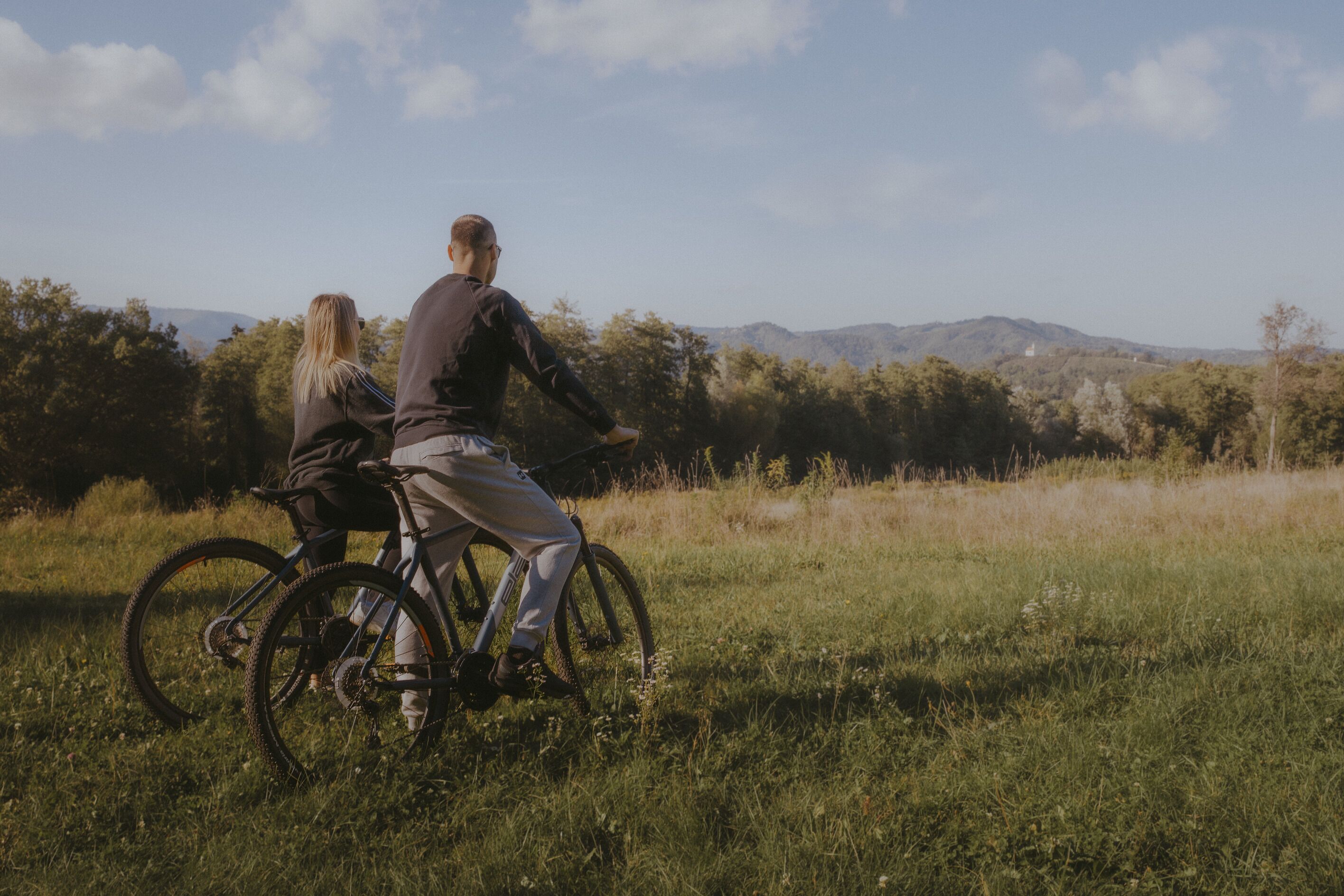 Two people riding bicycles on a grassy field with trees and mountains in the background.