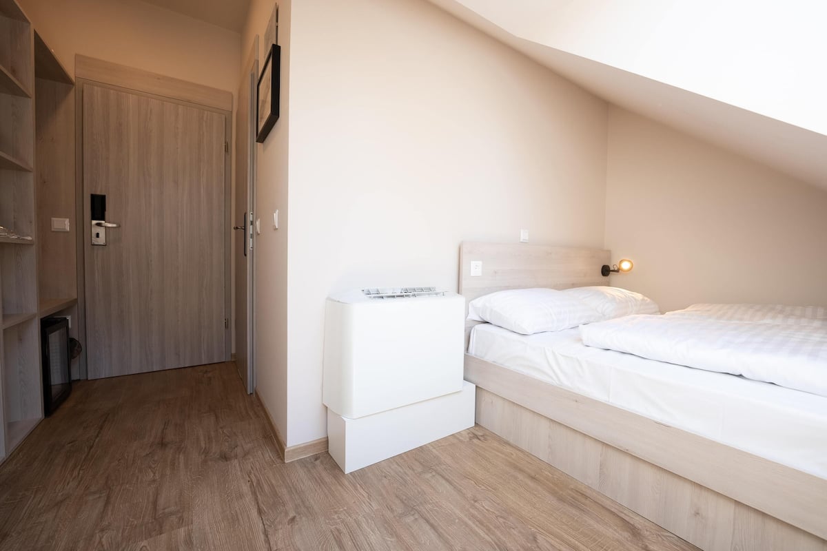 A bedroom with a wooden floor, a bed with white bedding, a nightstand, and a wall-mounted shelf.