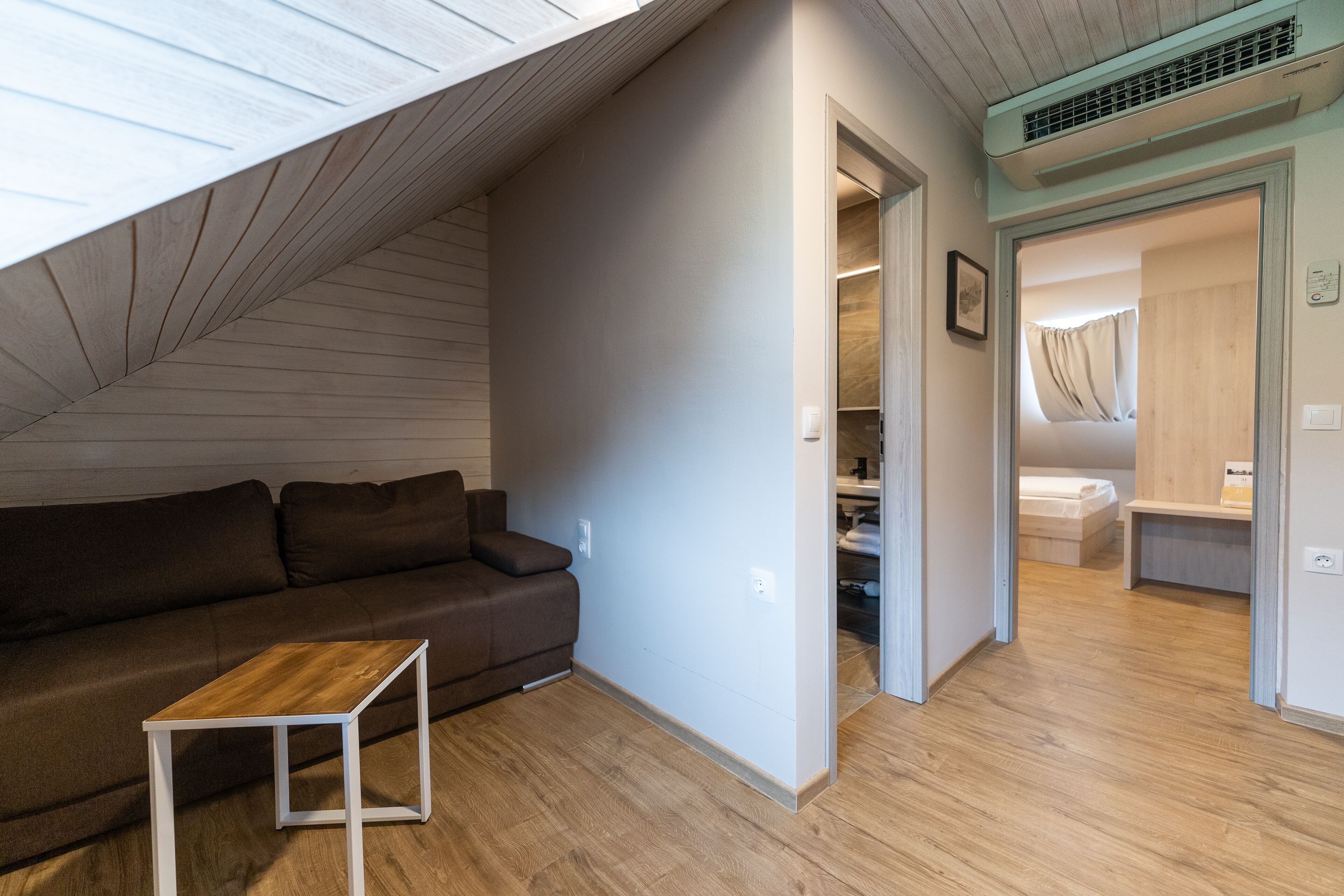 Un espace de vie moderne et compact, doté d’un plafond en pente, d’un plancher en bois, d’un canapé marron et d’une petite table blanche.