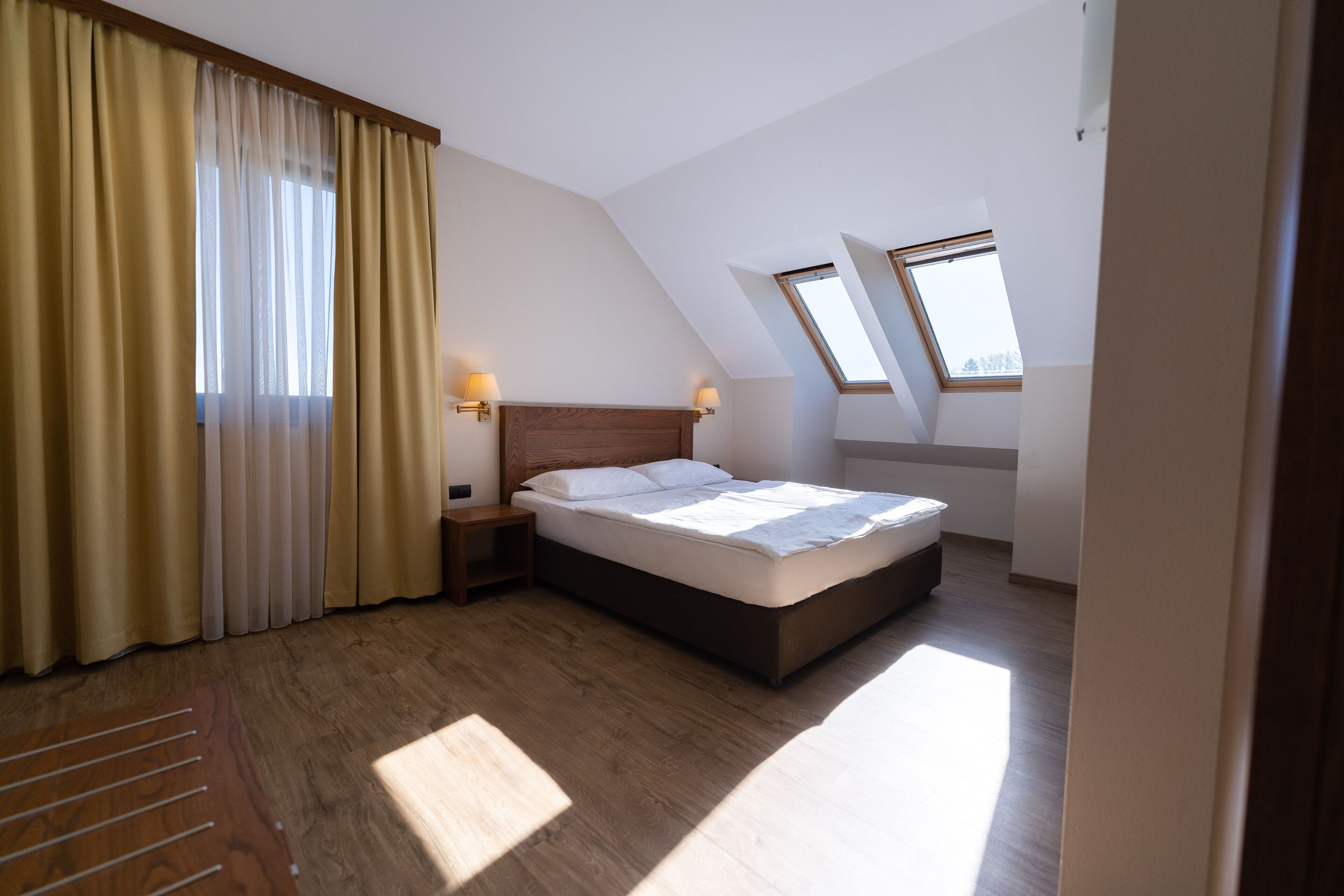 Un dormitorio con cama, piso de madera, una ventana con cortinas y un tragaluz.