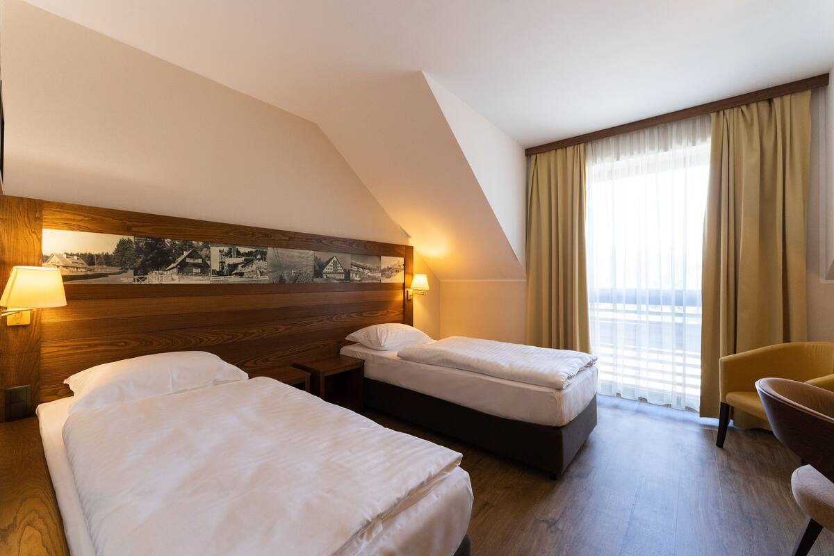 A hotel room with two beds, a wooden headboard, a painting, a chair, and a window with curtains.