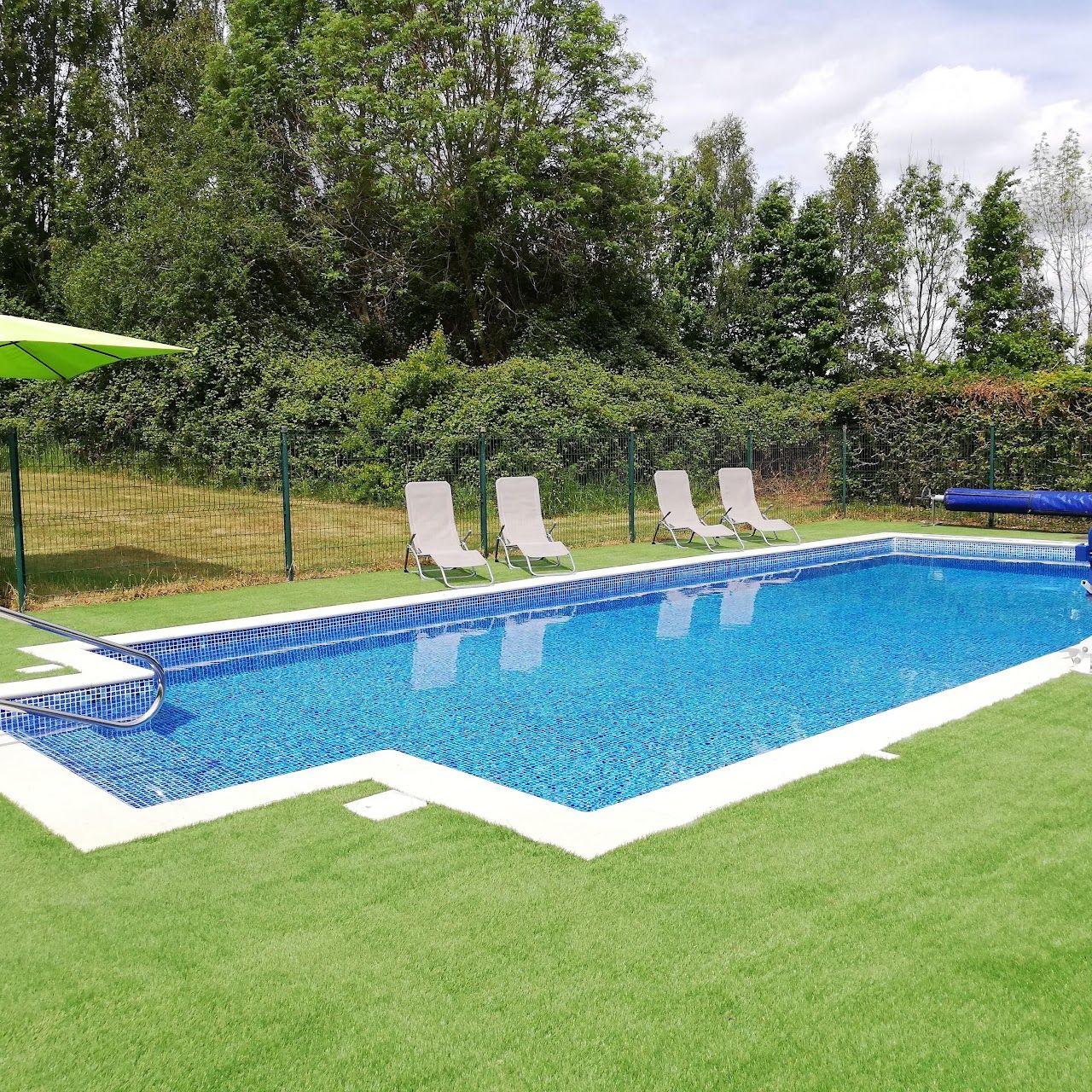 A rectangular swimming pool with a tiled edge, surrounded by a grassy area with white lounge chairs and a green umbrella.