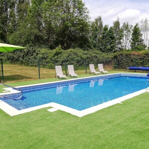 Une piscine rectangulaire dont les bords sont recouverts de carreaux, entourée d’une pelouse où se trouvent des chaises longues blanches et un parasol vert.
