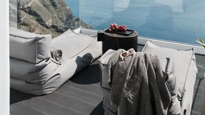 A balcony with a view of the sea, a glass table with a tray of grapes, and a cozy seating area with cushions and a blanket.