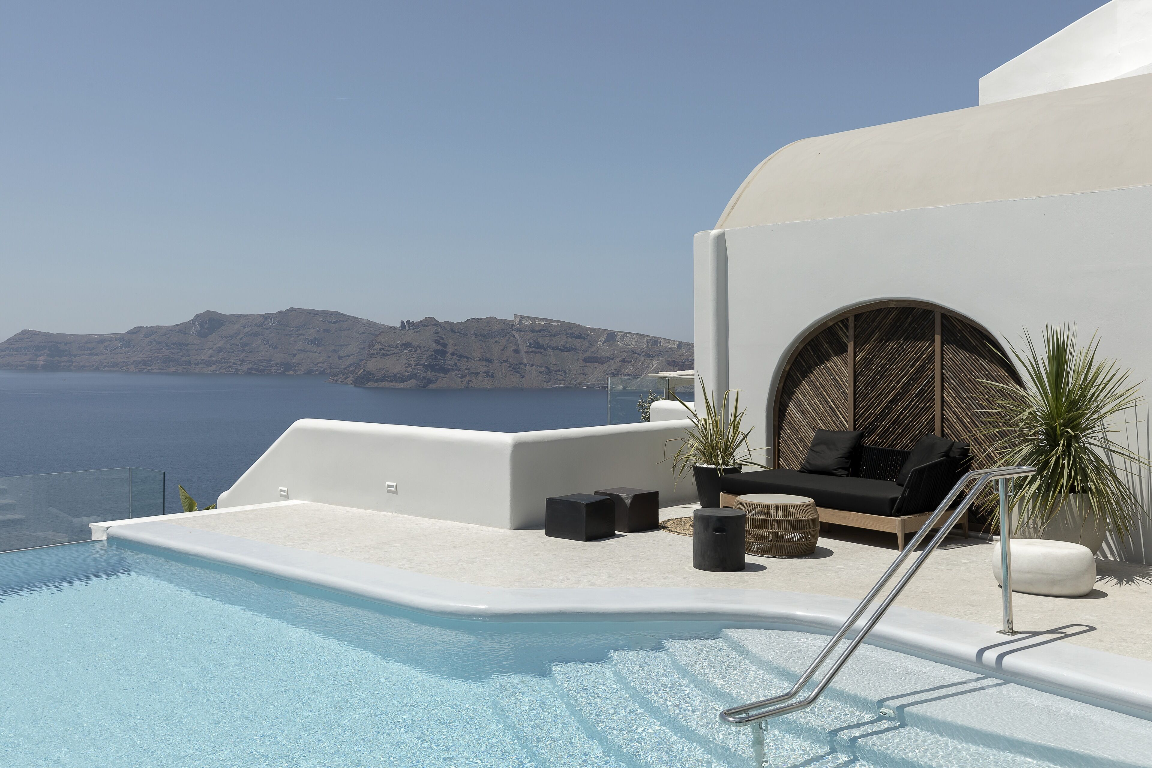 A rooftop pool area with a clear view of the sea and mountains, furnished with black sofas and potted plants.