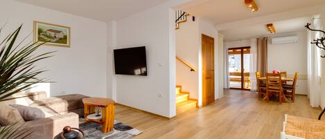 A modern living room with a sofa, a coffee table, and a TV mounted on the wall. There is a staircase leading to another room, and a dining area with a table and chairs.
