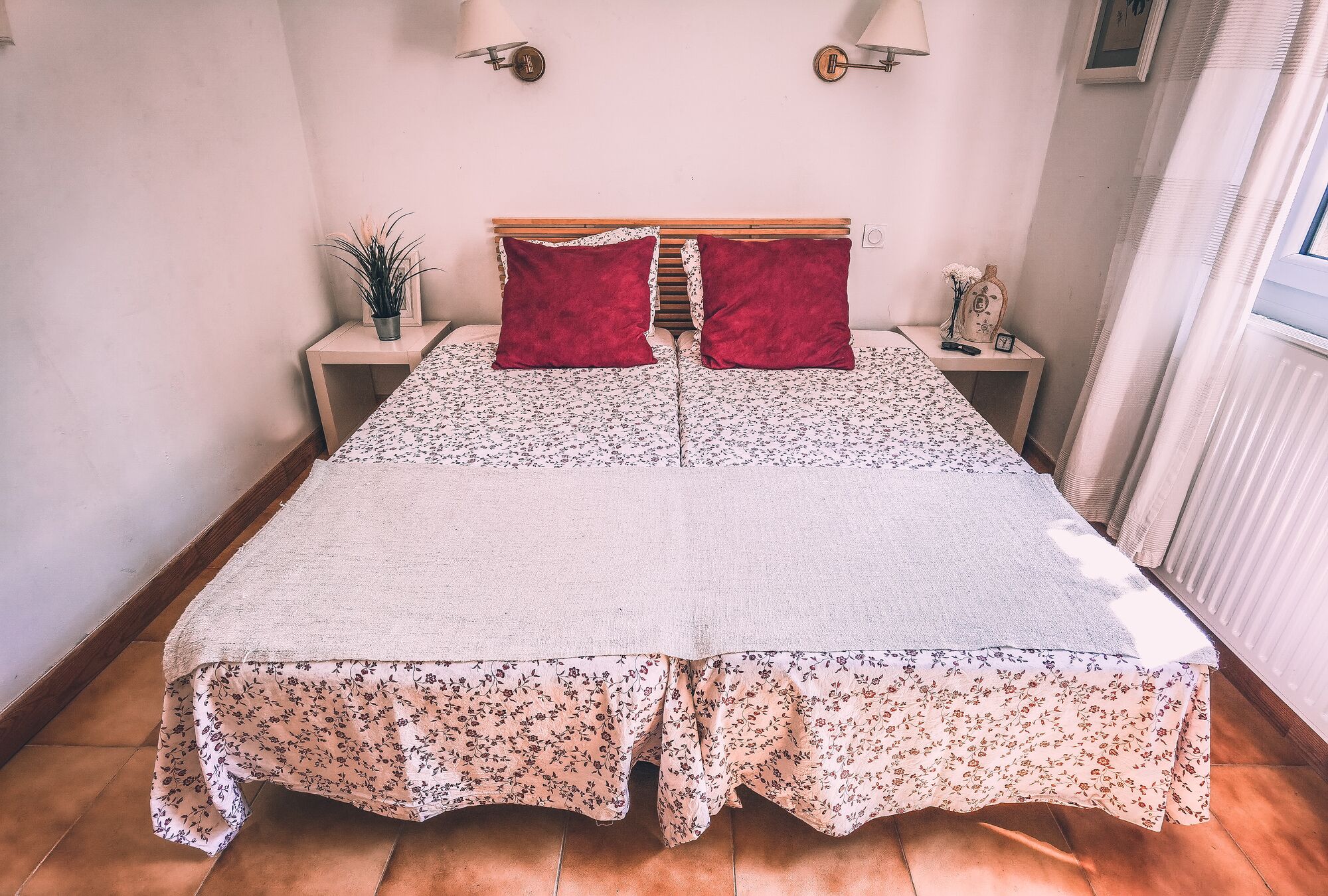 A double bed with floral bedding, two red pillows, a wooden headboard, bedside tables with lamps, and a potted plant.