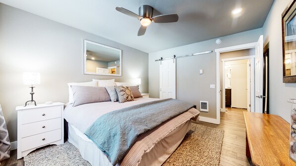 Un dormitorio con cama, mesitas de noche, ventilador de techo, espejo y puerta que da a otra habitación.