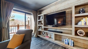 Un salon doté d’un grand t éléviseur à écran plat fixé à un mur en bois, d’un canapé confortable avec un coussin et d’une bibliothèque contenant divers objets.