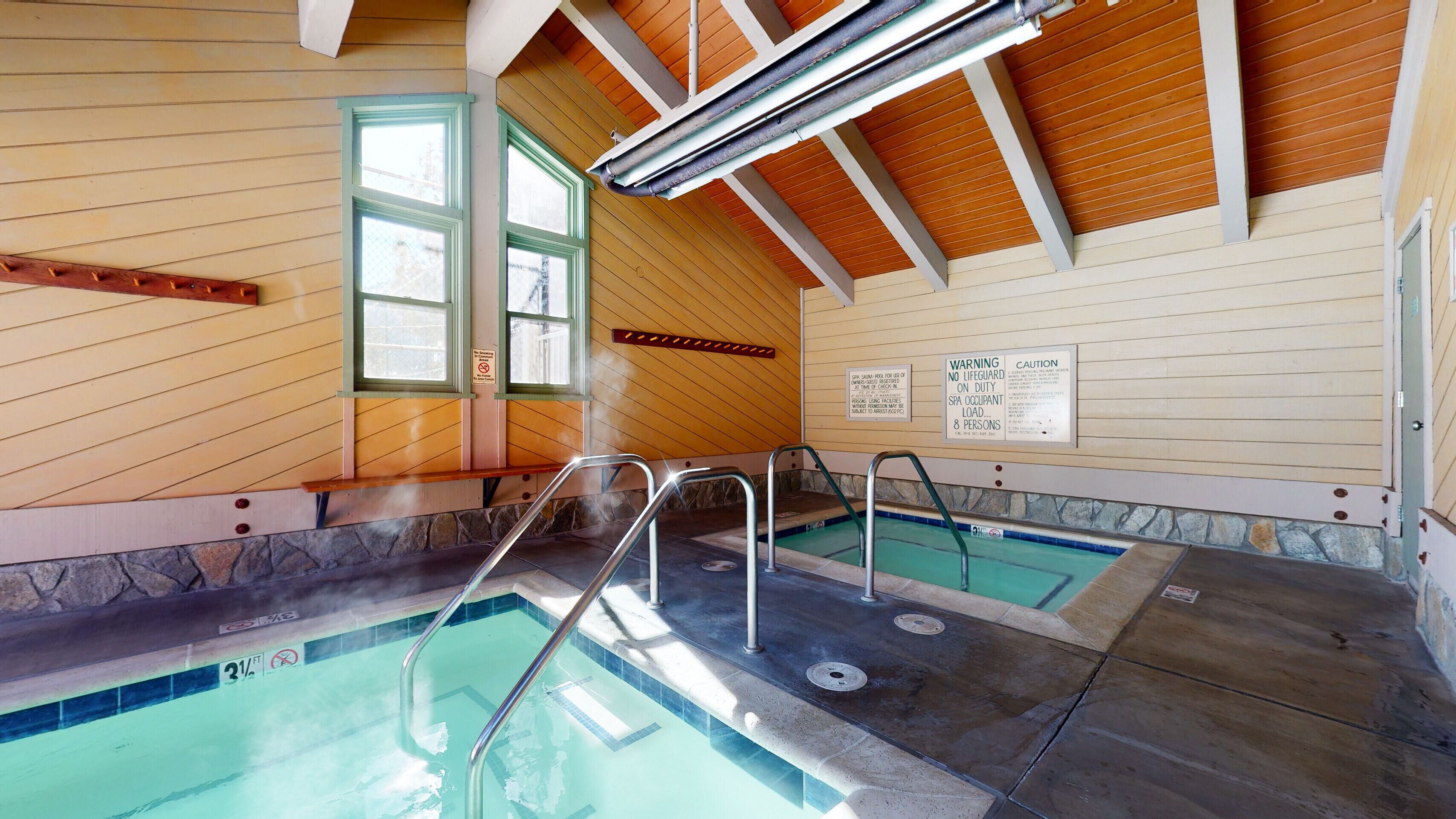 Zona d'spa amb banyera d'hidromassatge, piscina i sauna. Hi ha senyals que indiquen que no hi ha socorrista de servei i un límit de capacitat per a l'spa.