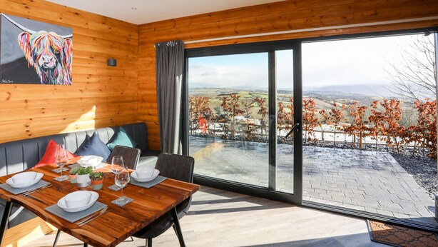 A modern dining area with a wooden table set for two, a sofa with cushions, and a large window offering a scenic view of the countryside. - Hilltop Lodge (Corwen)