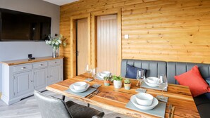 A dining area with a wooden table set for four, a grey sofa with a red pillow, and a grey cabinet with a flat-screen TV above it. - Hilltop Lodge (Corwen)