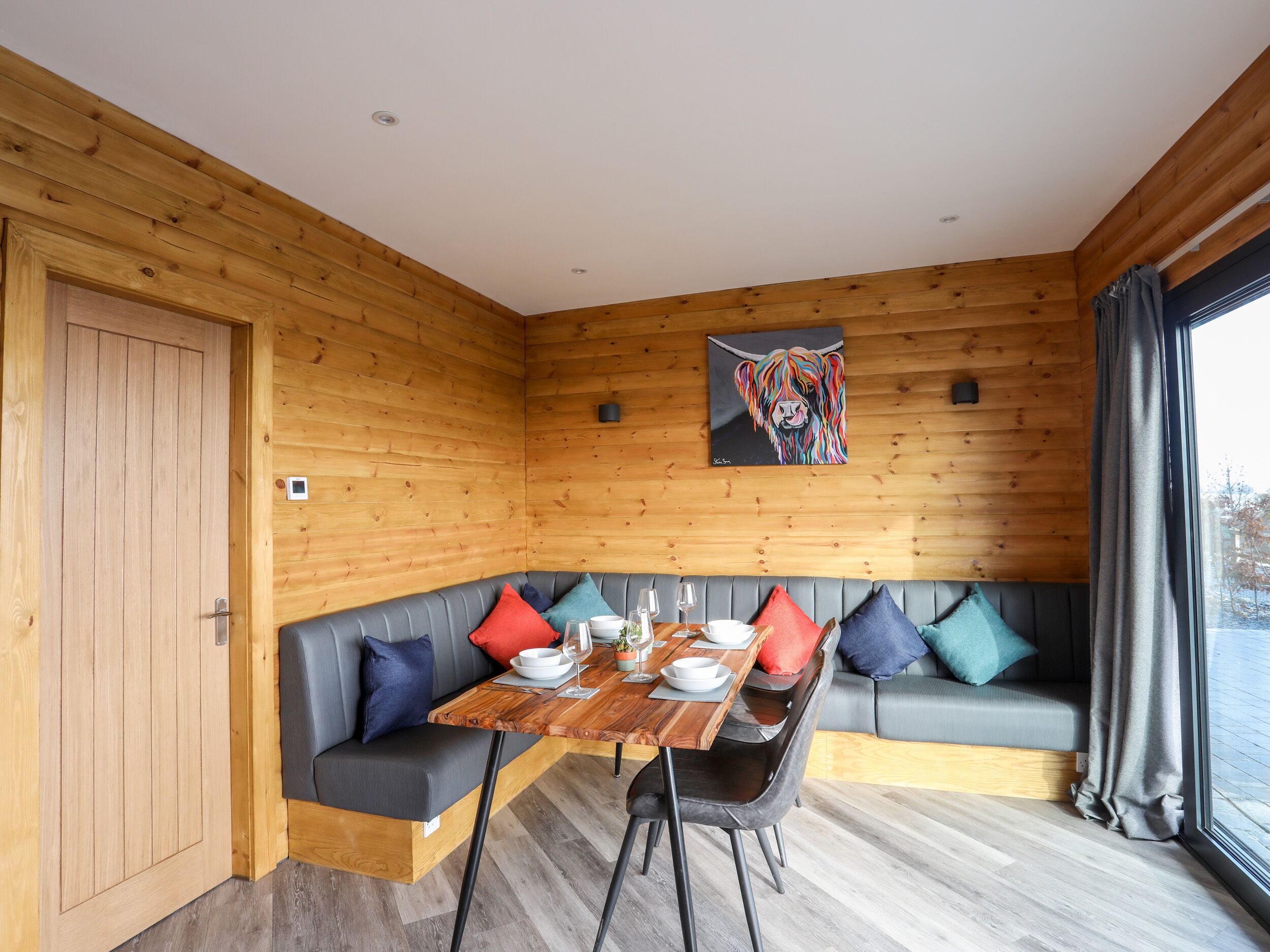 Een gezellig chaletinterieur met een houten wand, een eettafel met stoelen, een grijze bank met kleurrijke kussens en een schilderij aan de muur.