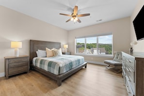 A bedroom with a large bed, two bedside tables with lamps, a ceiling fan, a window with a view, and a dresser.