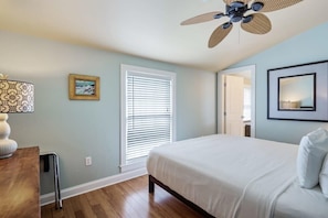 Ein Schlafzimmer mit einem Bett, einem Deckenventilator, einem Fenster mit Jalousien, einer Lampe und einem Bild an der Wand.