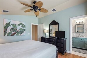 Un dormitorio con cama, cómoda, ventilador de techo, un cuadro y un baño con lavamanos y espejo.
