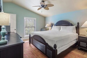A bedroom with a bed, bedside tables, a ceiling fan, a window with blinds, and a door.