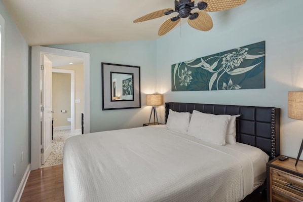 A bedroom with a large bed, a ceiling fan, a framed floral artwork, and a bedside lamp.