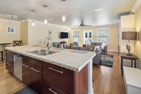 A spacious kitchen with a large island, a dining area with a table and chairs, a living room with a sofa and a TV, and a ceiling fan.